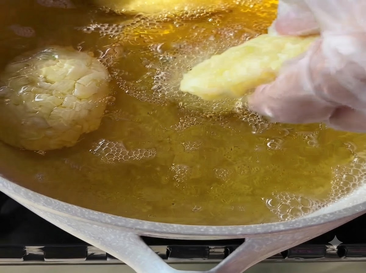 Una mano con guante colocando suavemente una croqueta de papa moldeada en una sartén con aceite de cocina caliente burbujeando.