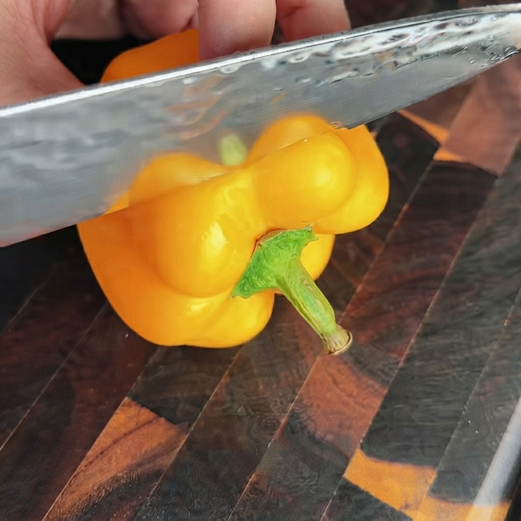 Un cuchillo afilado cortando la sección superior de un pimiento amarillo brillante en una tabla de cortar de madera.