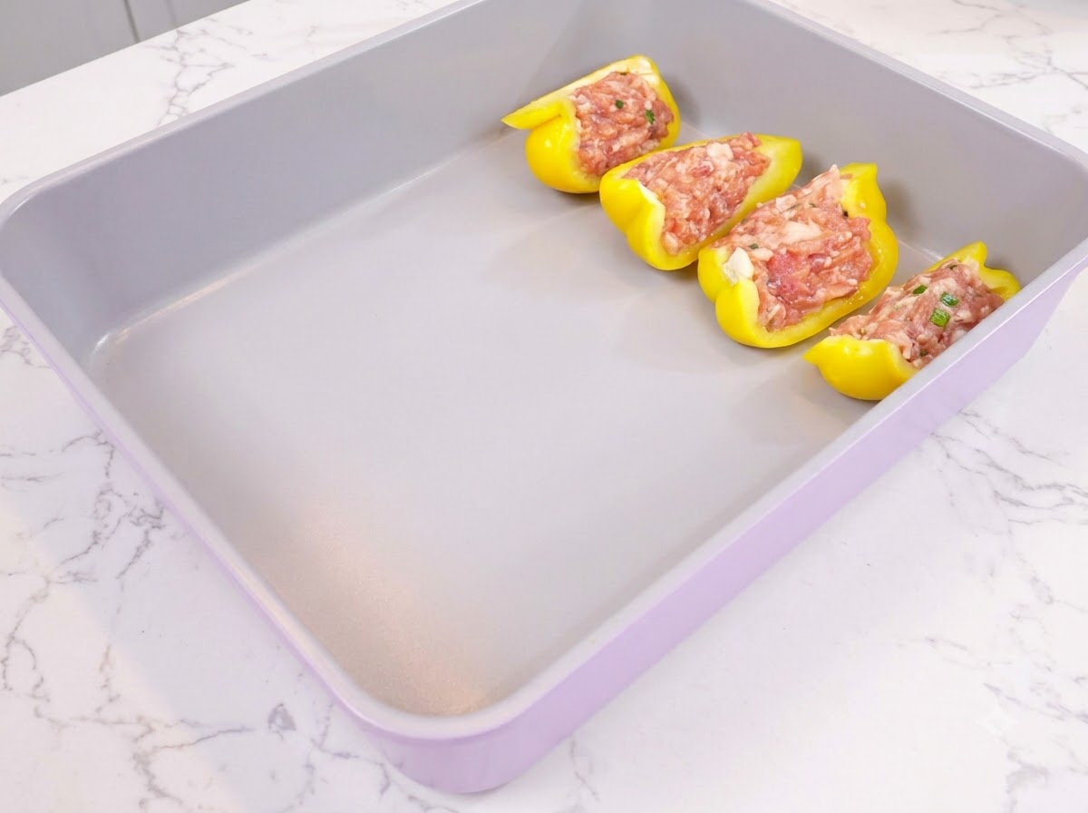 Mitades de pimiento amarillo rellenas de carne picada dispuestas ordenadamente en una bandeja de horno morada.