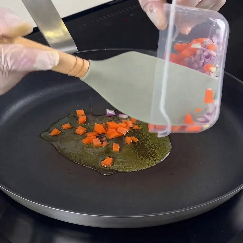 Cebollas, zanahorias, ajos y hierbas picadas siendo transferidos a una sartén caliente con aceite.