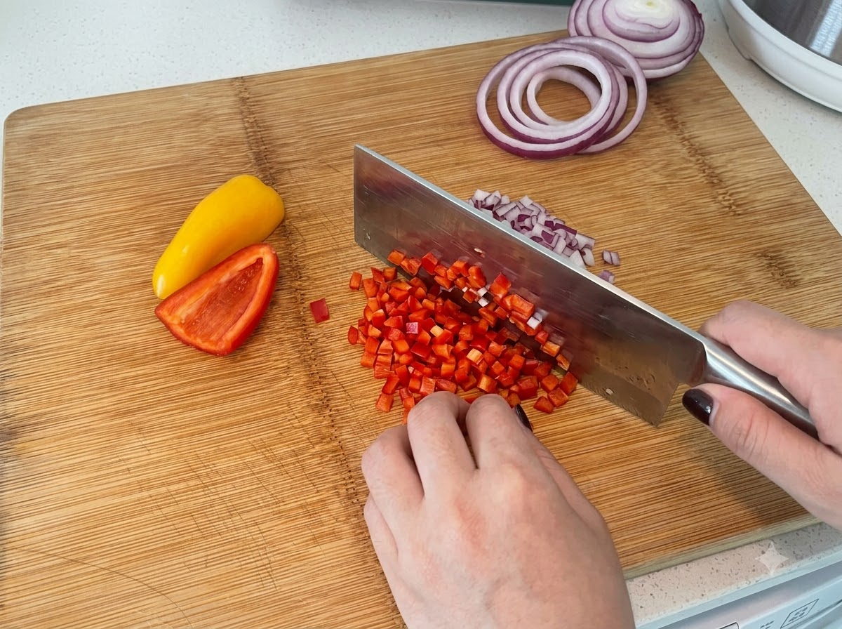 Pimientos rojos y amarillos siendo cortados en cubitos sobre una tabla de cortar de madera con un cuchillo de cocina.