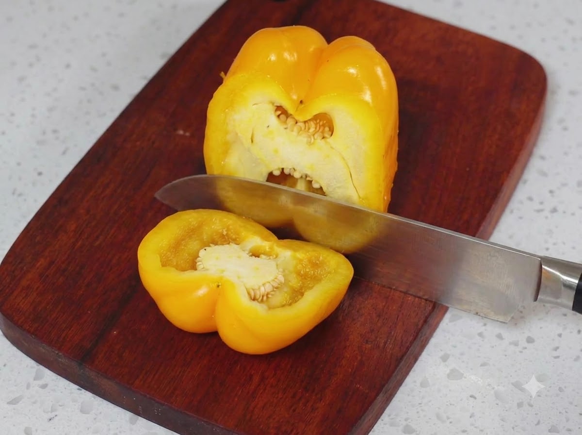 Un pimiento amarillo siendo cortado por la mitad en una tabla de cortar de madera con un cuchillo de cocina.