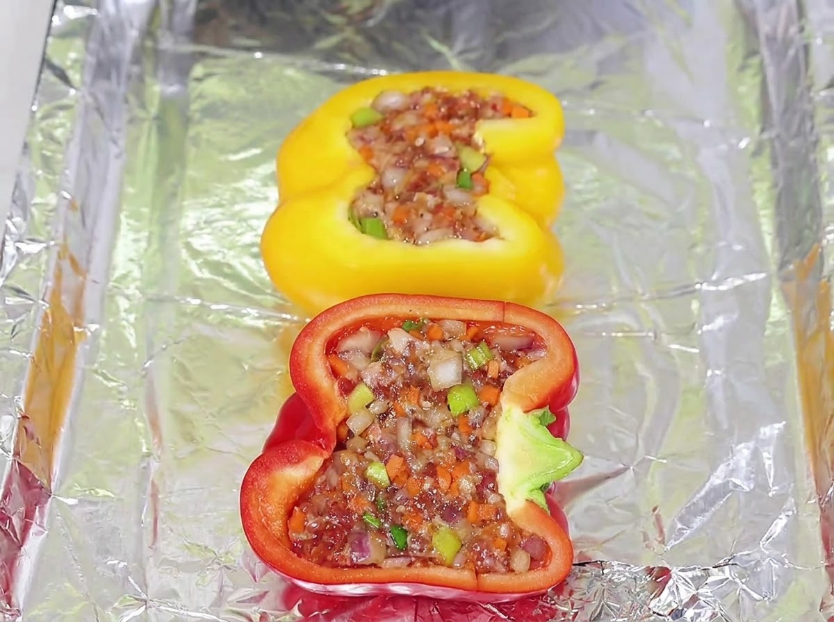 Mitades de pimiento amarillo y rojo rellenas con mezcla de carne dispuestas ordenadamente en una bandeja de horno forrada con papel de aluminio.