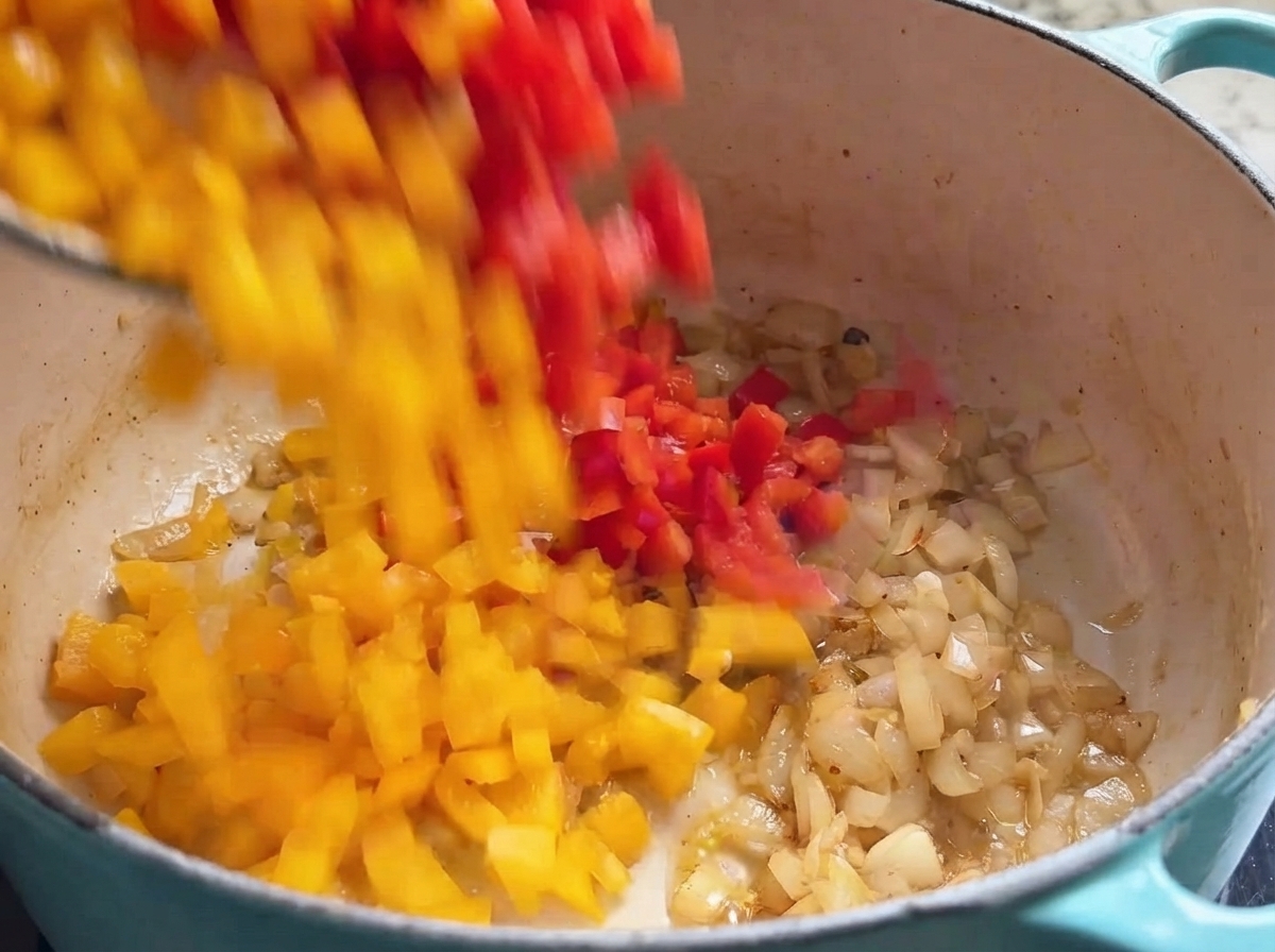 Dados de pimiento rojo y amarillo cayendo en una olla de cocción llena de cebollas sofritas.