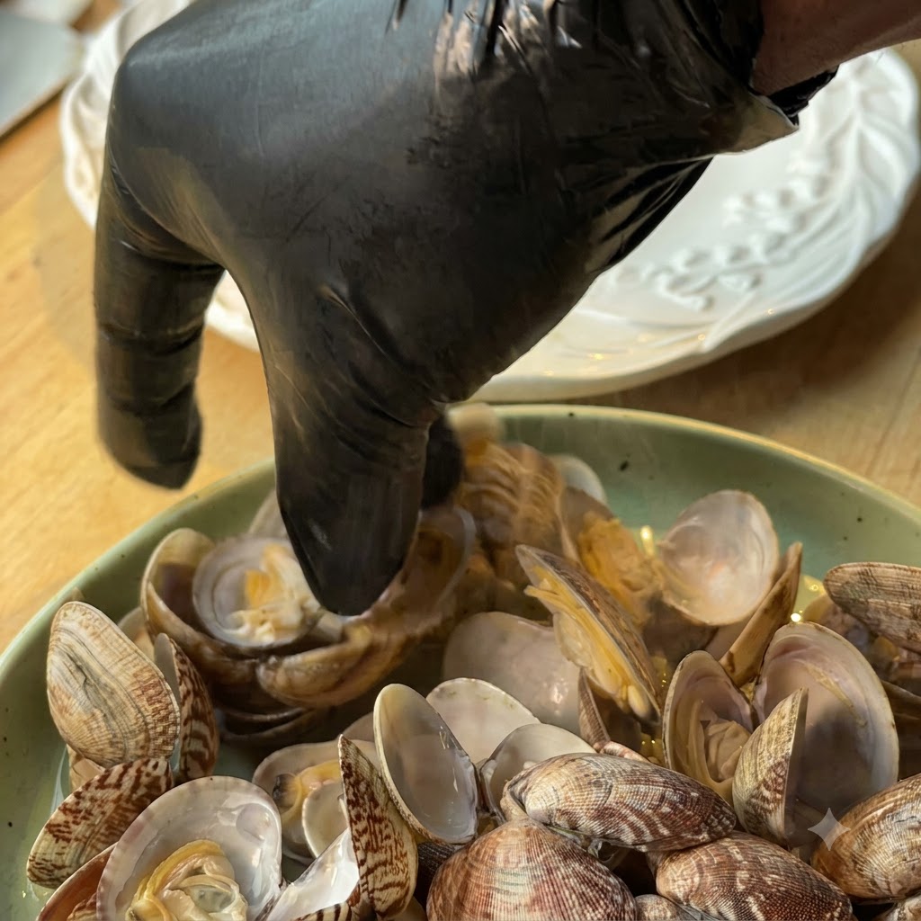 Una persona con guantes negros sacando la carne de almeja de las conchas hacia un plato.