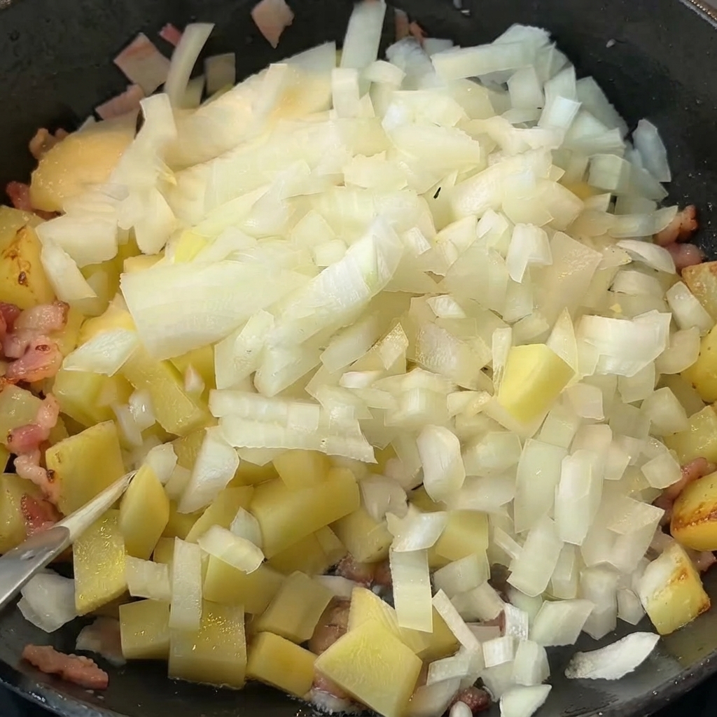 Cebollas picadas siendo vertidas sobre papas cocidas y tocino en una sartén.