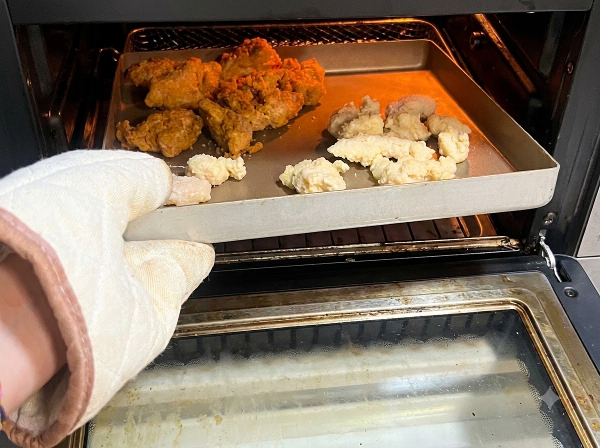 Un guante de horno sosteniendo una bandeja llena de popcorn chicken y alitas picantes crudas o congeladas dentro de una freidora de aire.