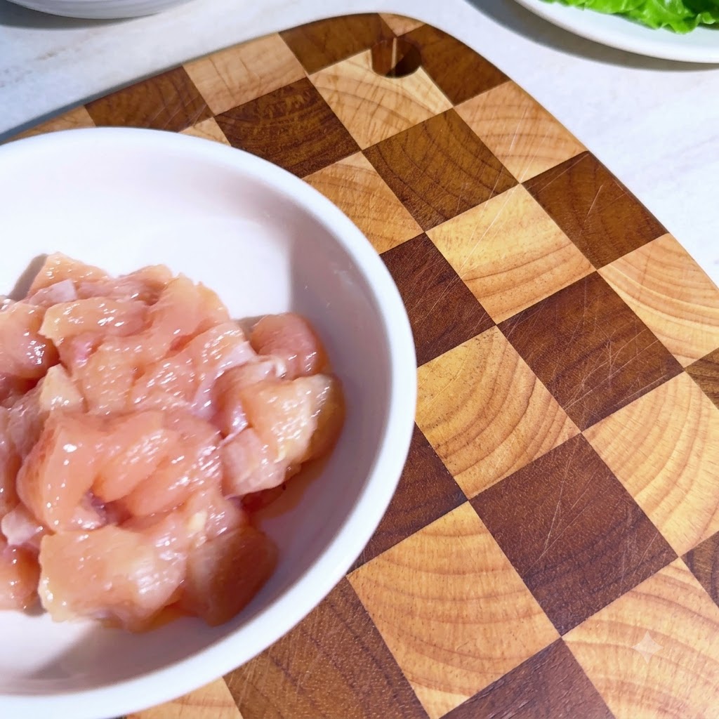 Pequeños trozos de pollo en un tazón blanco con especias medidas espolvoreadas encima antes de mezclar.