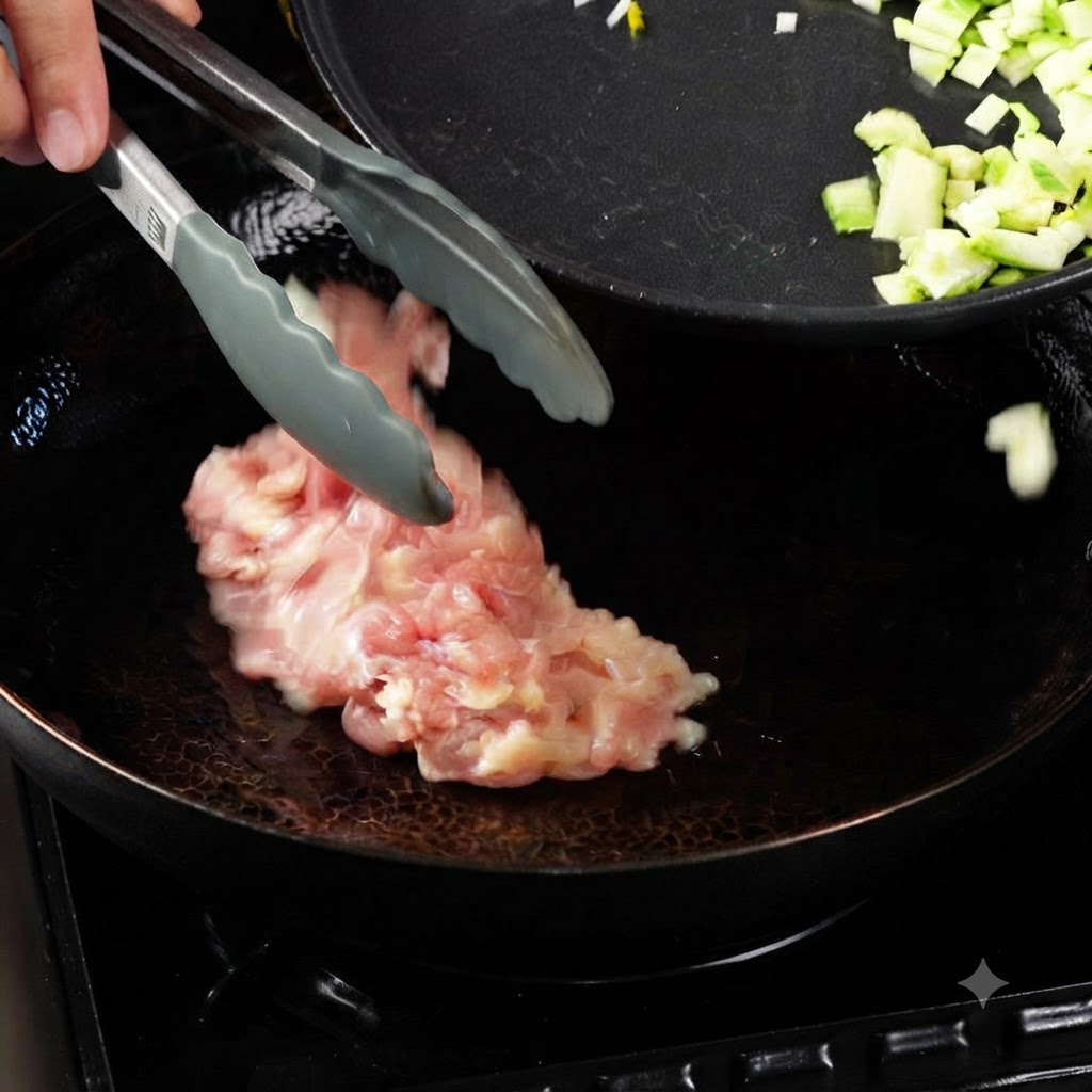 Pinzas colocando trozos de pollo crudo en un wok caliente con aceite, junto a una sartén con verduras cortadas en daditos.