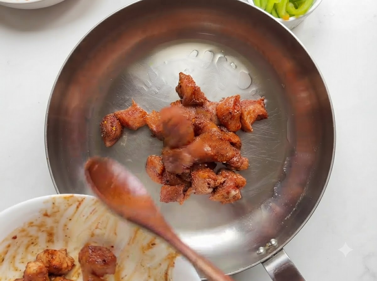 Trozos de pollo marinado cocinándose y dorándose en una sartén de acero inoxidable.
