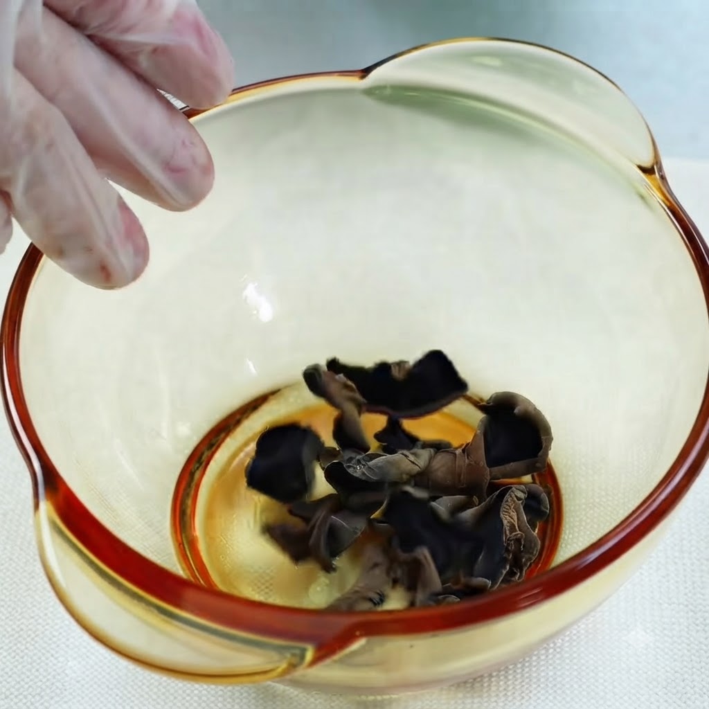 Agua vertiéndose desde una taza medidora de vidrio en un tazón con hongos oreja de madera negros secos.