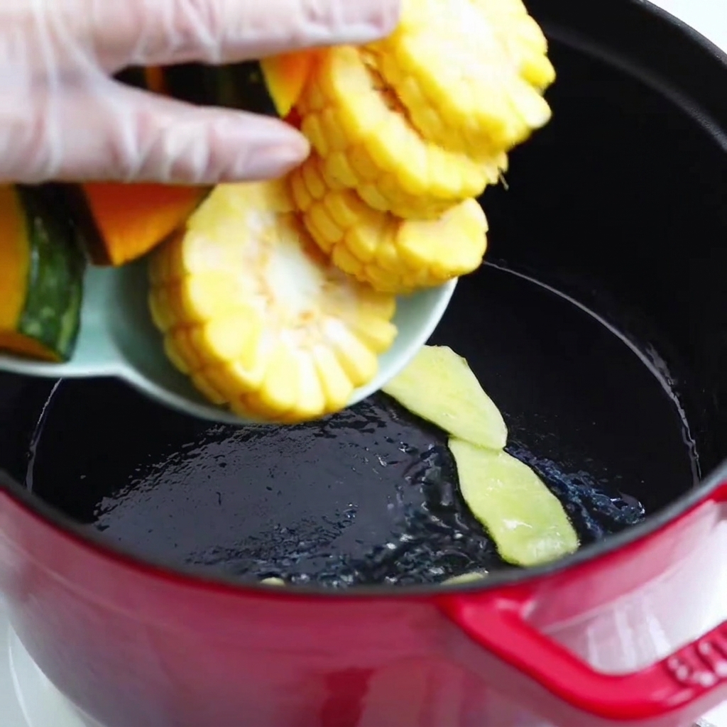 Trozos de calabaza y mazorcas de maíz vertidos desde un plato a la olla roja.