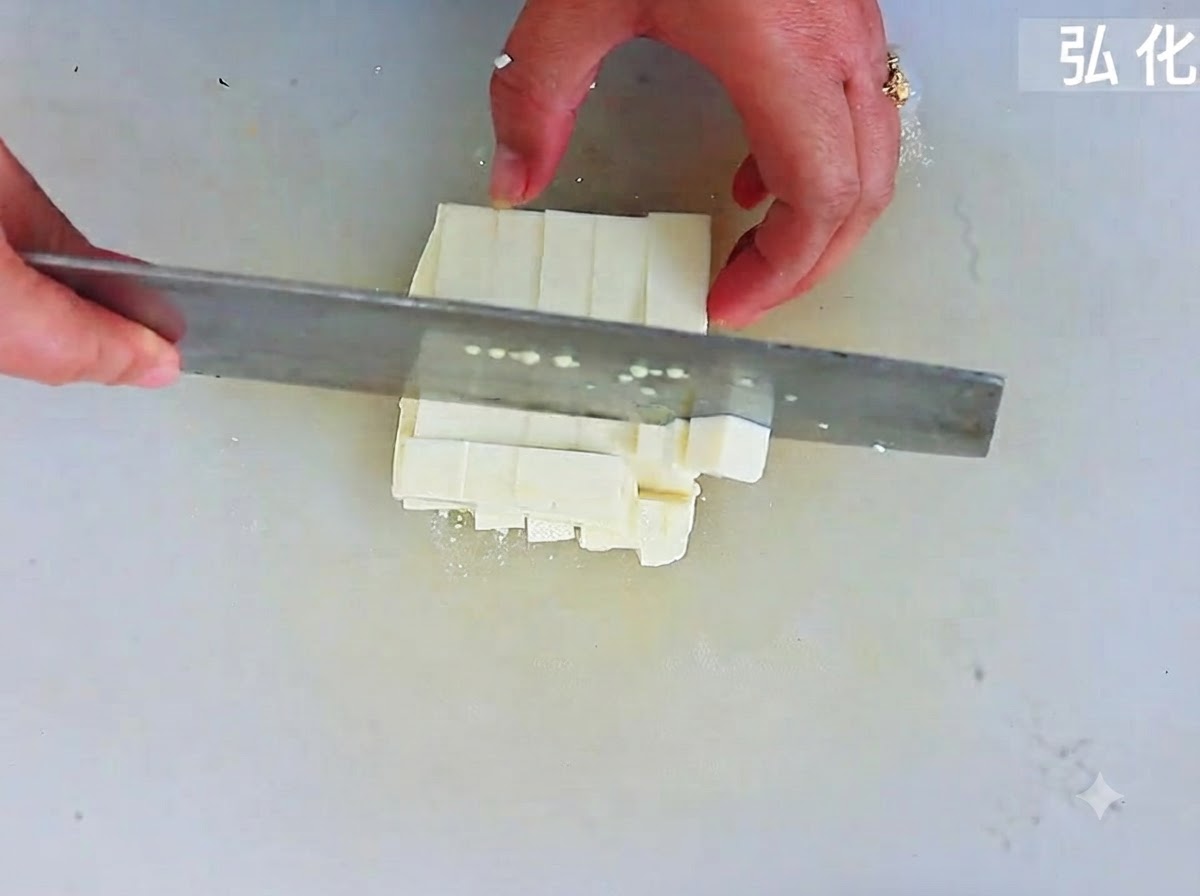 Tofu tierno siendo cortado en cubos cuadrados uniformes con un cuchillo grande sobre una tabla de cortar.