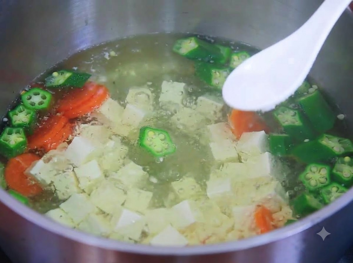 Una cuchara blanca sosteniendo sal sobre una olla humeante de sopa de tofu, okra y zanahoria.