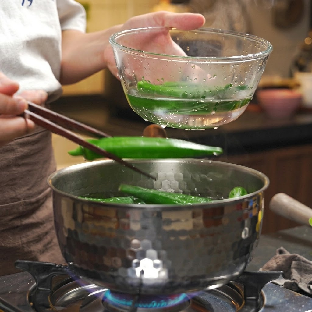 Okra cocida siendo transferida con palillos desde una olla caliente a un tazón transparente con agua helada.