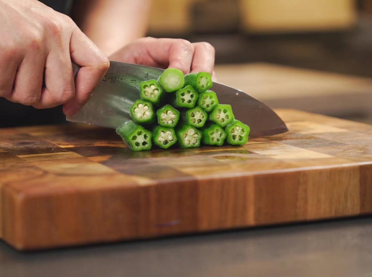 Un cuchillo de chef cortando los tallos y partiendo por la mitad las vainas de okra cocidas apiladas en una tabla de cortar.