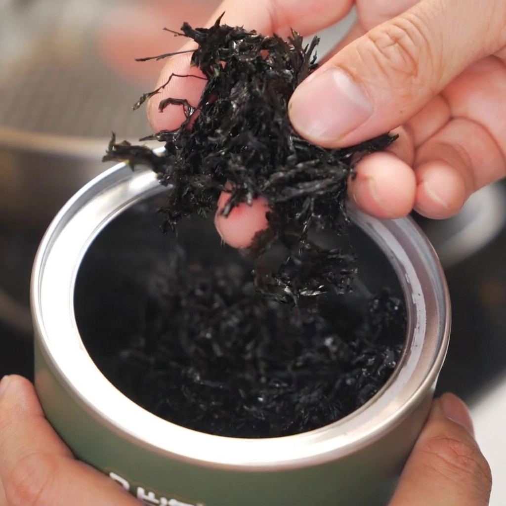 Manos sosteniendo alga seca y desmenuzada sobre una sartén de acero inoxidable caliente.