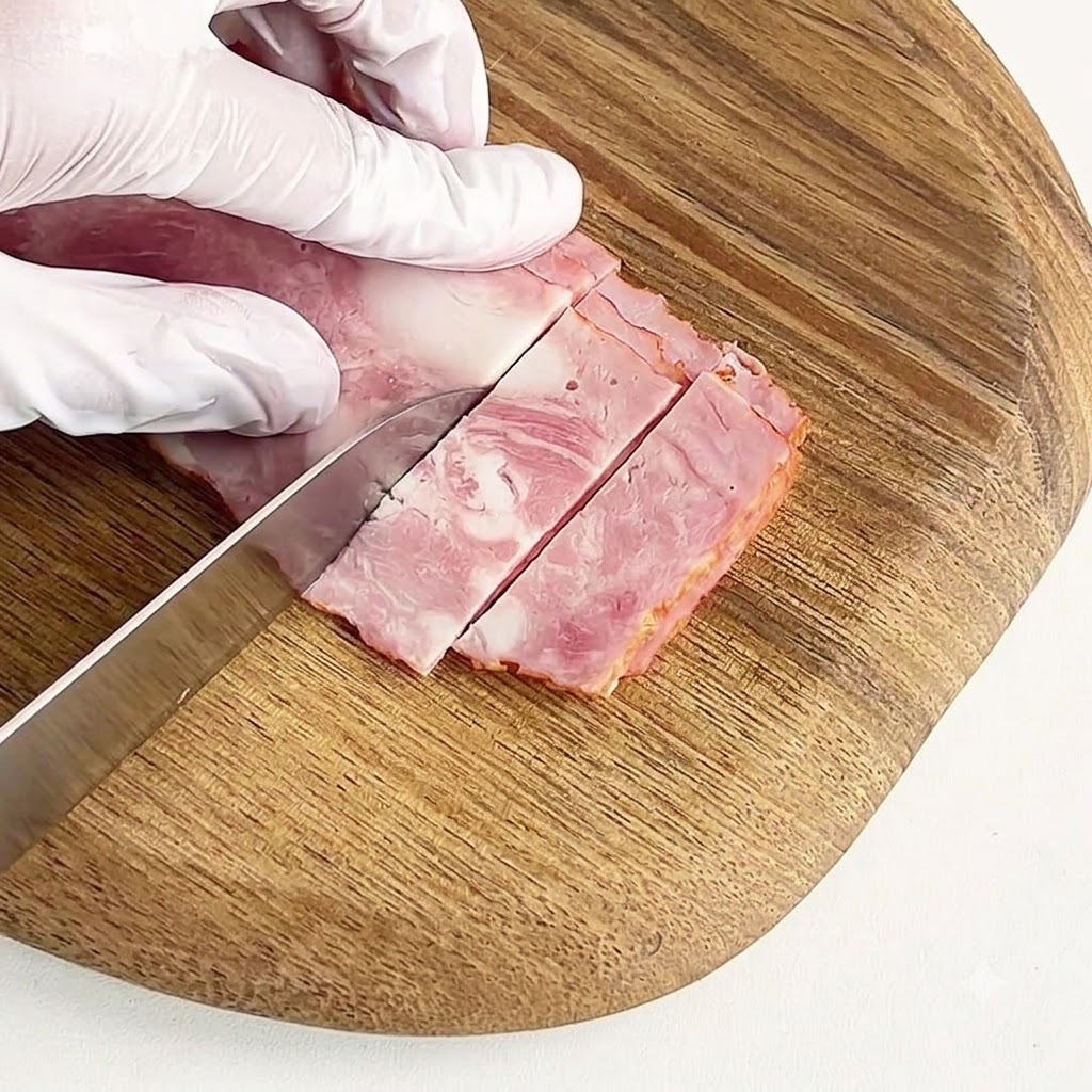 Una mano con guante cortando tocino curado en trozos pequeños sobre una tabla de cortar de madera redonda.