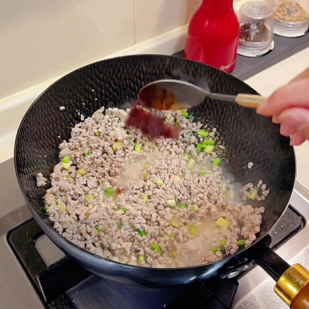 Una cucharada de doubanjiang siendo añadida a un wok que contiene carne picada y aromáticos.