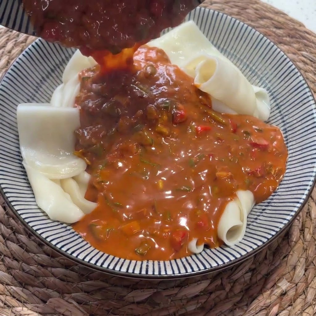 Salsa roja rica vertida desde un cucharón sobre un tazón lleno de fideos anchos doblados.