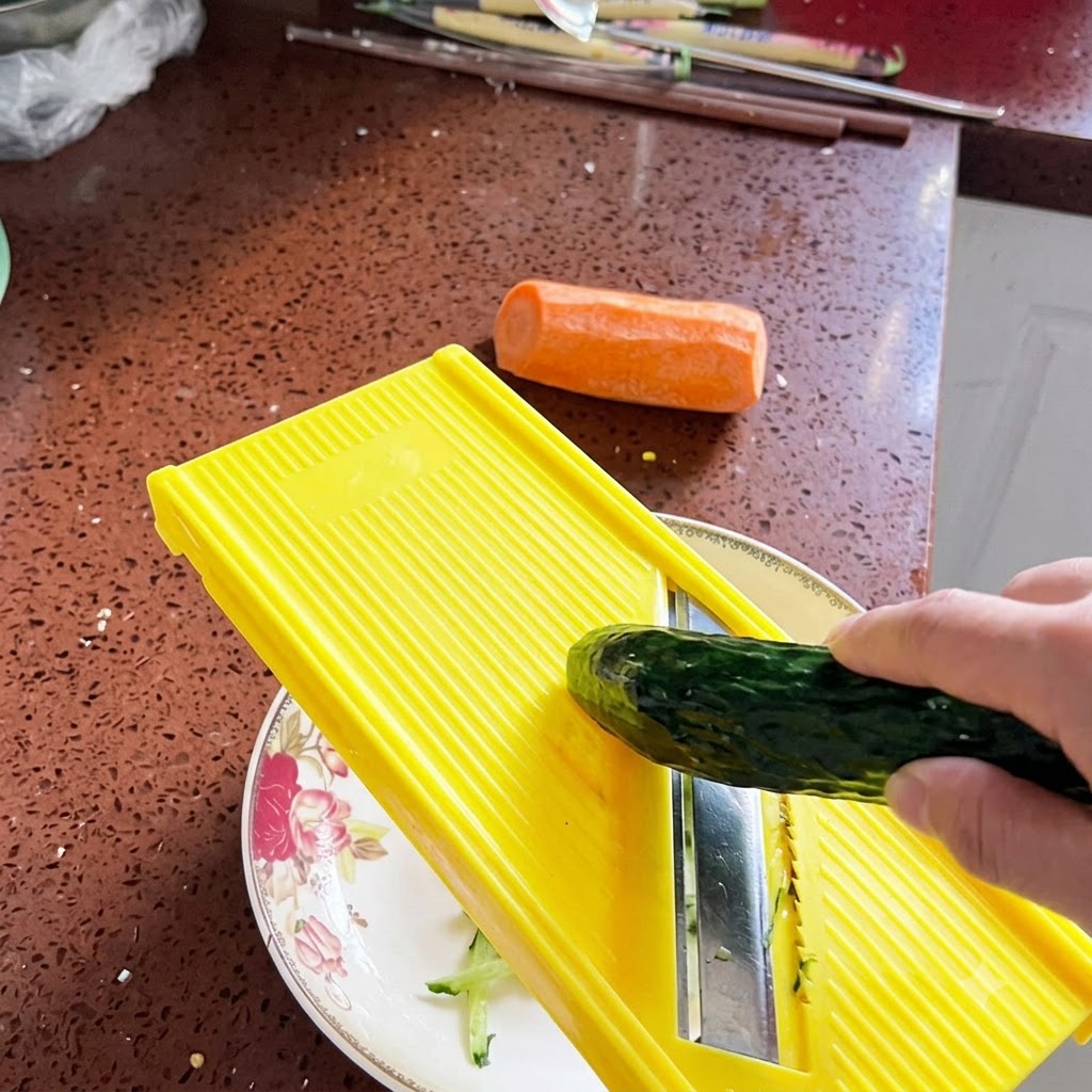 Un pepino fresco siendo rallado en tiras finas usando una mandolina de color amarillo brillante sobre un plato decorativo.
