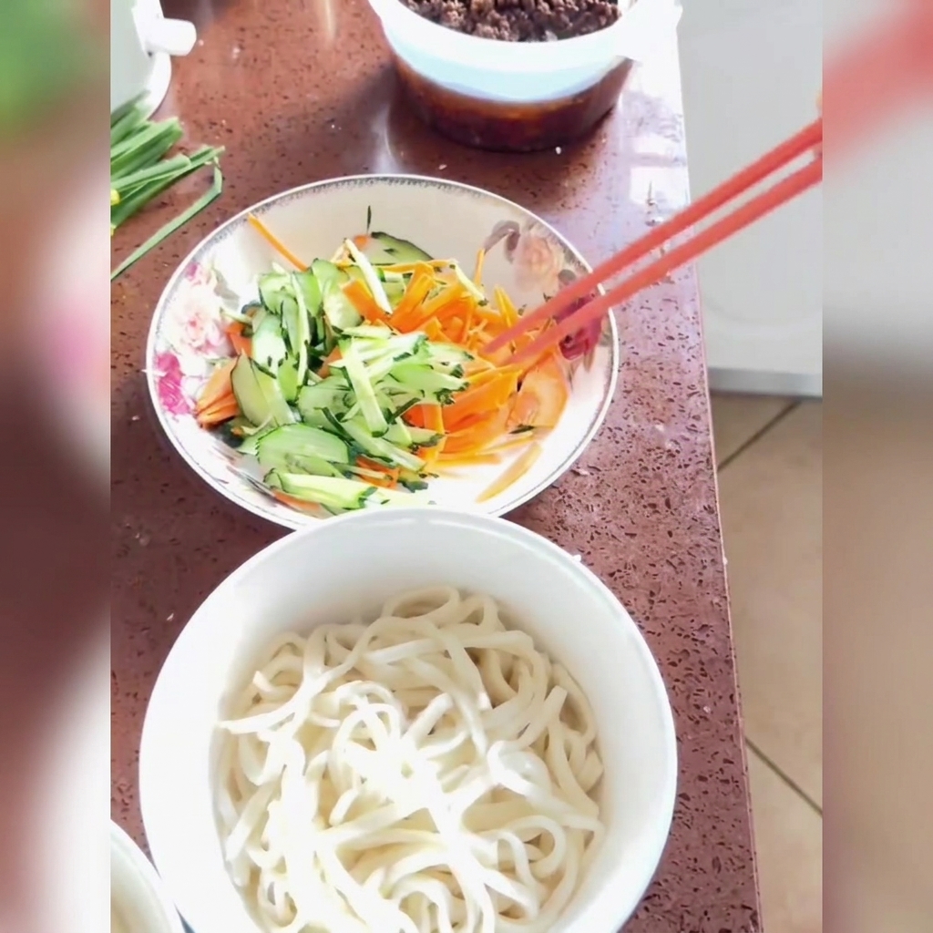 Salsa frita siendo servida y rociada sobre fideos recién cocinados cubiertos con pepino rallado y zanahorias en un tazón blanco.