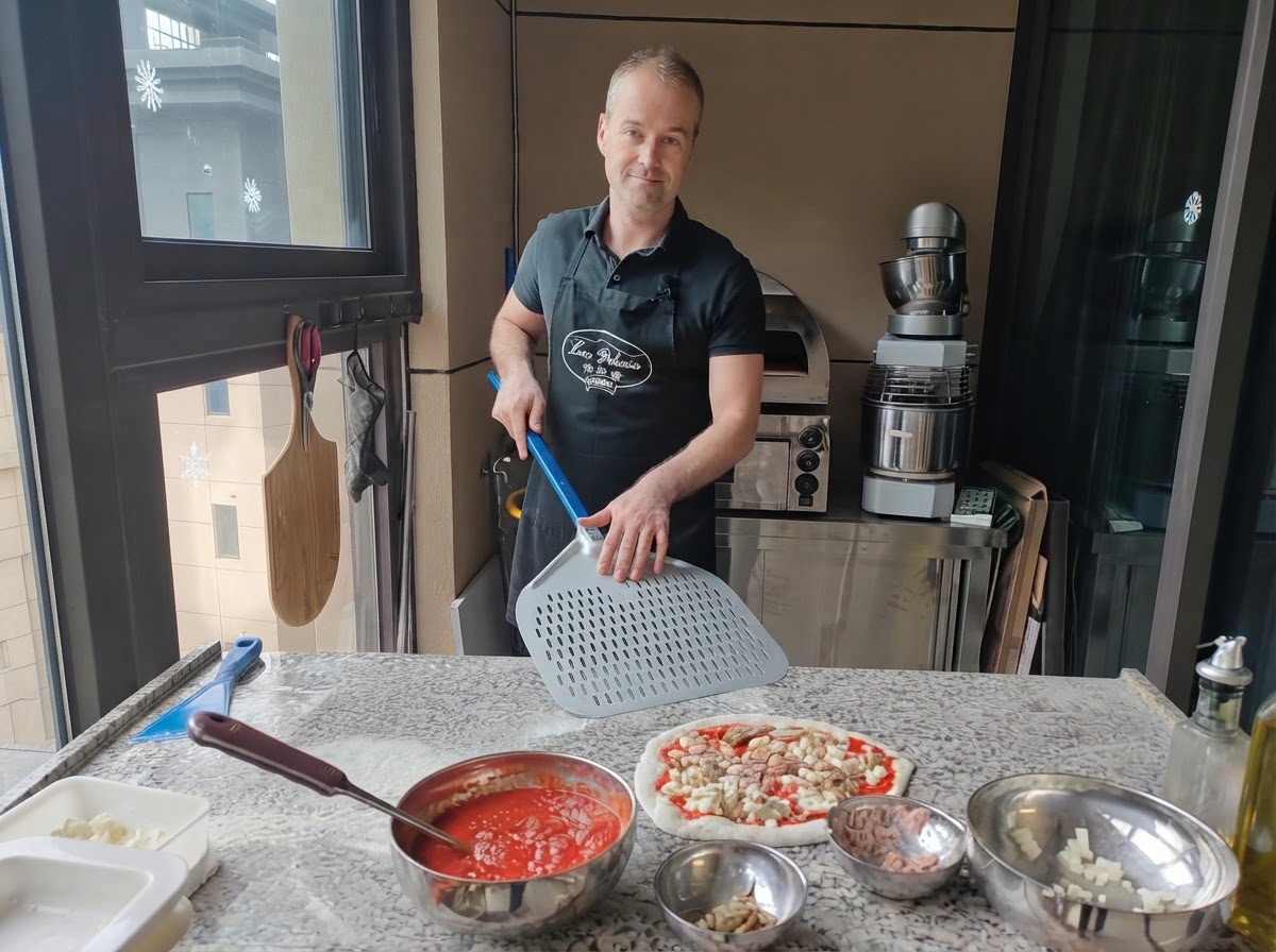 Un pizzero sosteniendo una gran pala de metal perforada posicionada justo al lado de una pizza cruda completamente armada.
