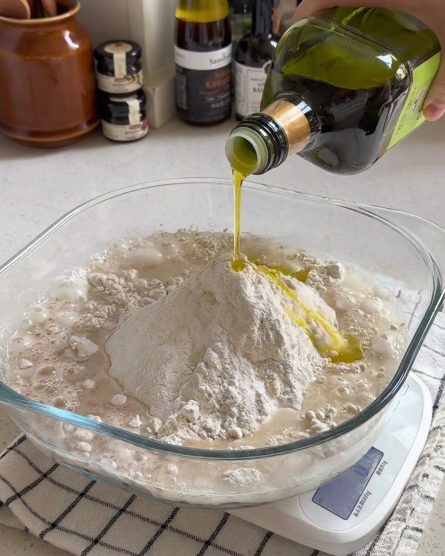 Aceite de oliva siendo vertido sobre una mezcla de harina y agua dentro de un recipiente de vidrio transparente en una balanza de cocina.