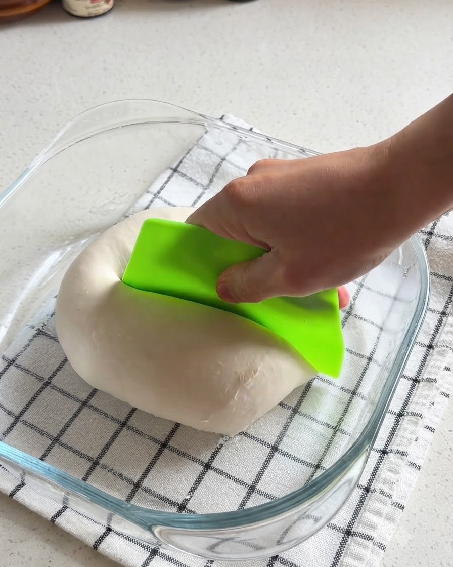 Una mano usando una rasqueta de banco de color verde brillante para cortar a la mitad una bola de masa de pizza lisa dentro de un recipiente de vidrio.