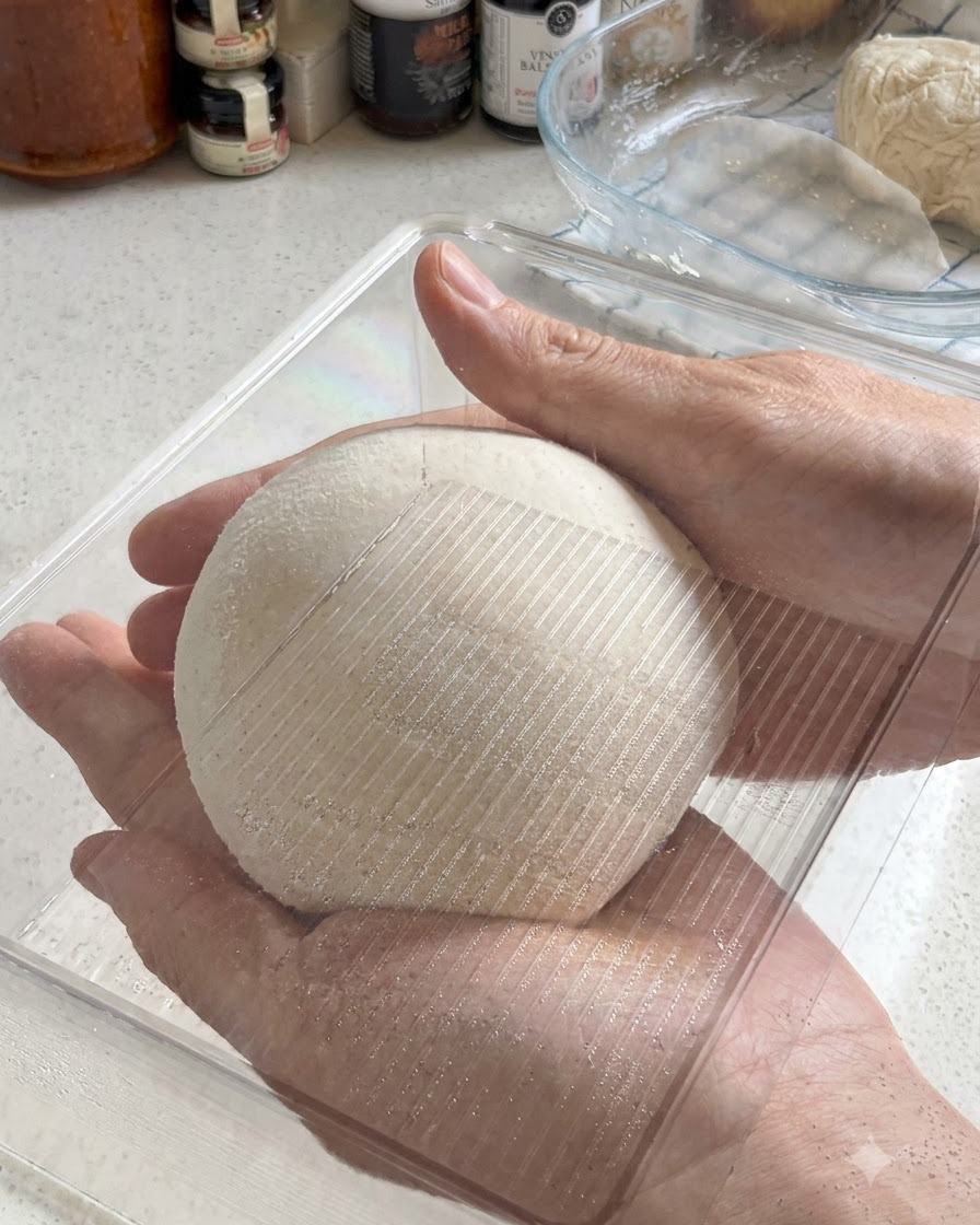 Dos manos sosteniendo una bola de masa de pizza perfectamente redondeada mientras se coloca en un recipiente de fermentación de plástico transparente.
