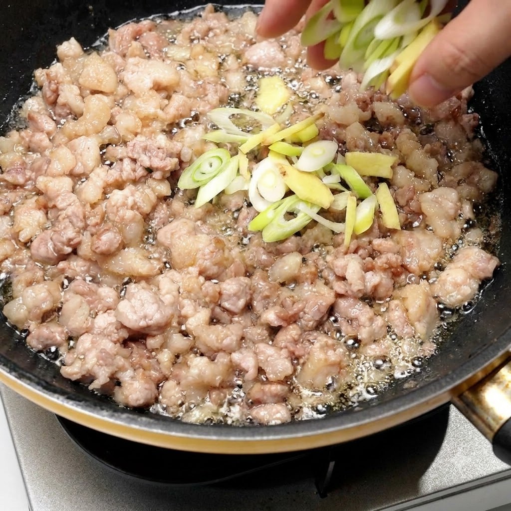 Cebolleta y jengibre picados siendo añadidos directamente a la sartén con la carne de cerdo dorada.