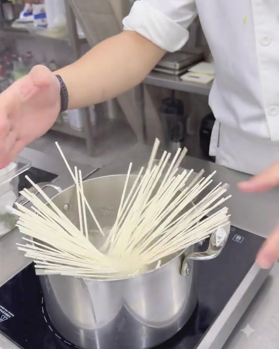 Fideos udon blancos secos cayendo en una olla de acero inoxidable con agua hirviendo.