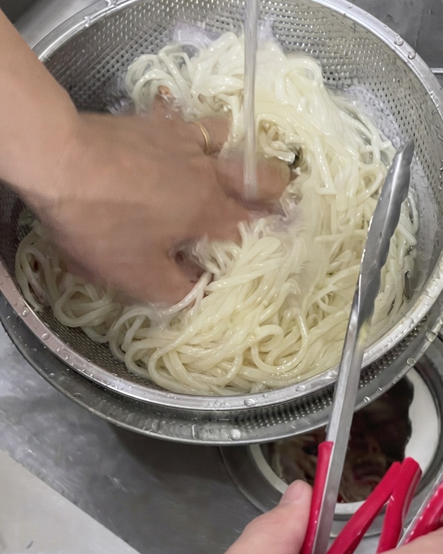 Una mano escurriendo fideos udon cocidos en un colador de metal bajo el agua fría del grifo.