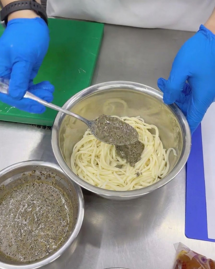 Manos con guantes usando una cuchara para añadir salsa espesa de trufa negra a los fideos udon fríos en un bol de metal.