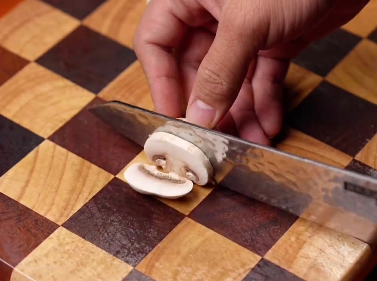 Champiñones siendo cortados con un cuchillo sobre una tabla de madera y luego añadidos a una sartén negra.
