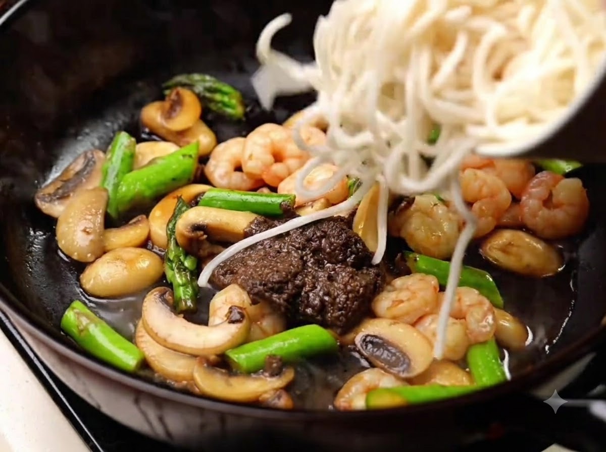 Fideos cocidos siendo añadidos a la sartén para mezclarse con la salsa, los champiñones, los camarones y los espárragos.