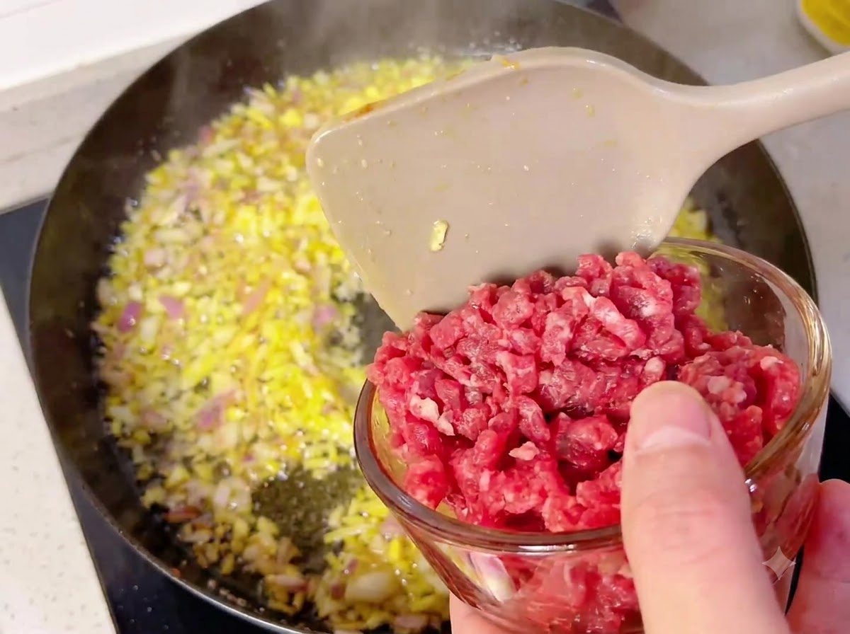 Un tazón pequeño de vidrio vertiendo cubos de carne en una sartén caliente llena de cebollas y ajo salteados.