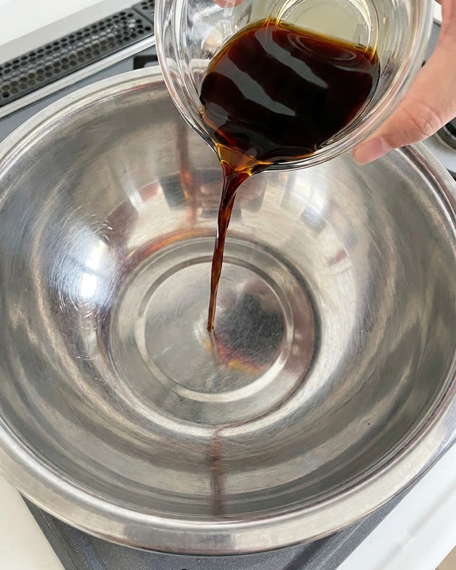 Salsa de soja oscura siendo vertida desde un recipiente de vidrio en un tazón de acero inoxidable.
