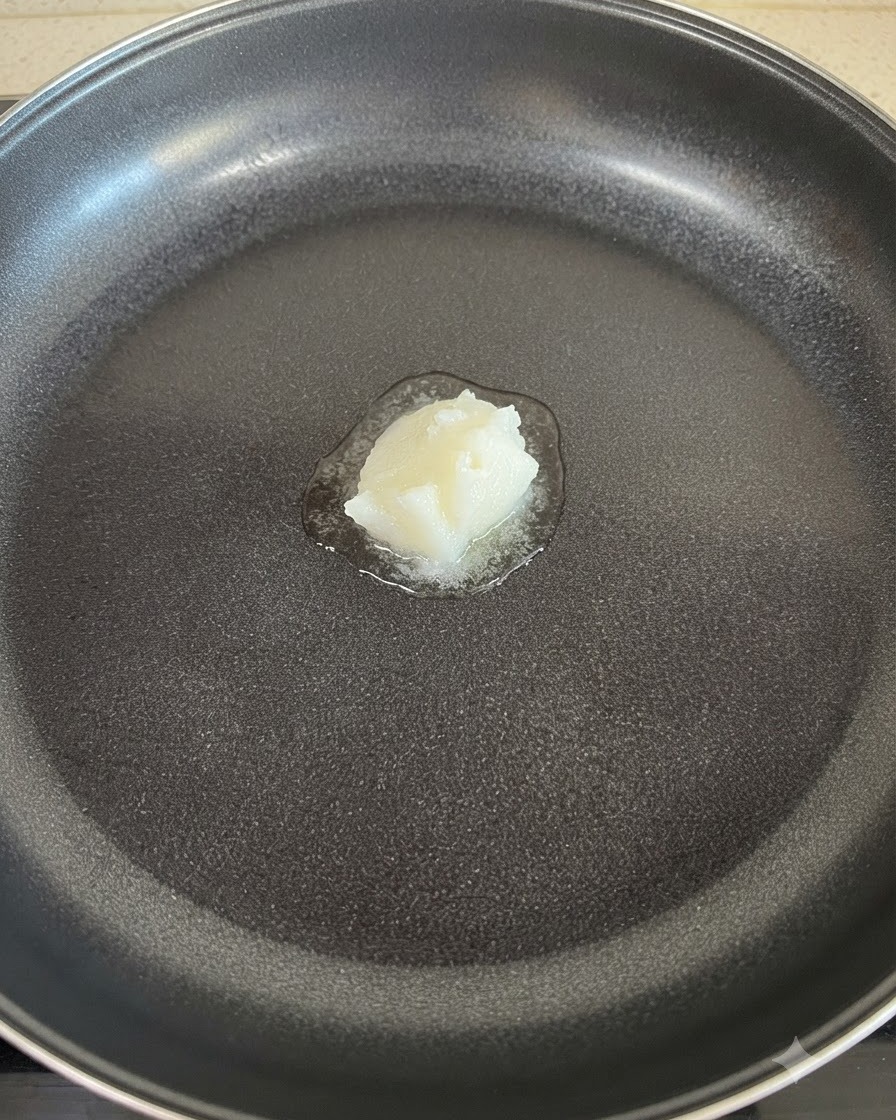 Una porción sólida de manteca blanca derritiéndose en el centro de una sartén antiadherente.