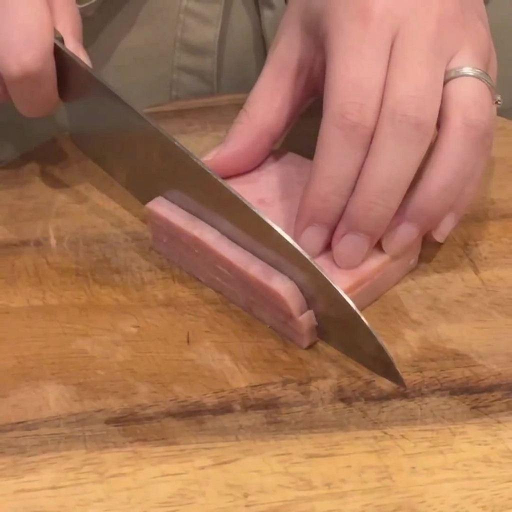 Un cuchillo de cocina cortando un bloque de jamón rosado en tiras finas y uniformes sobre una tabla de madera.