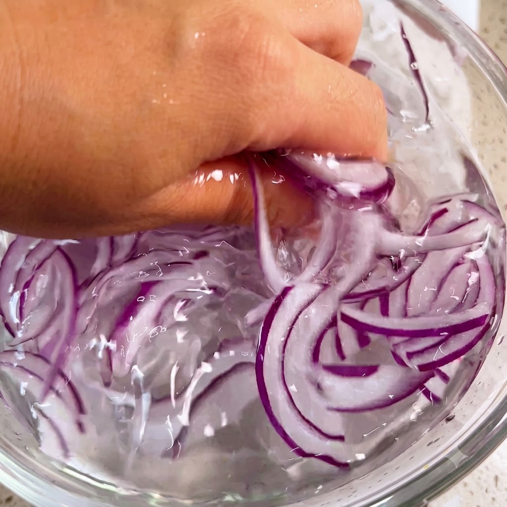 Una mano lavando suavemente tiras de cebolla morada dentro de un bol lleno de agua con hielo.