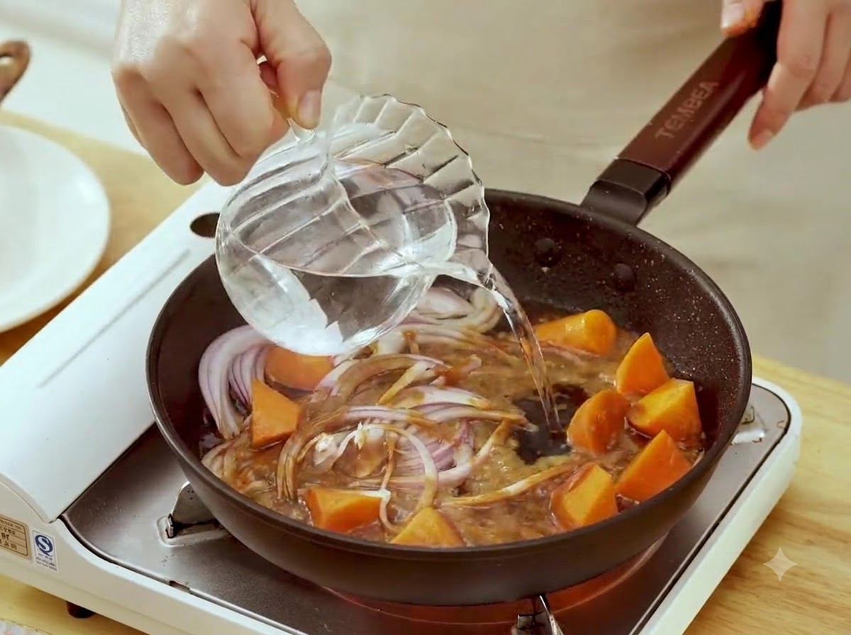 Agua siendo vertida desde una jarra de cristal en una sartén con cebollas rojas cortadas y trozos de zanahoria en salsa de miso.