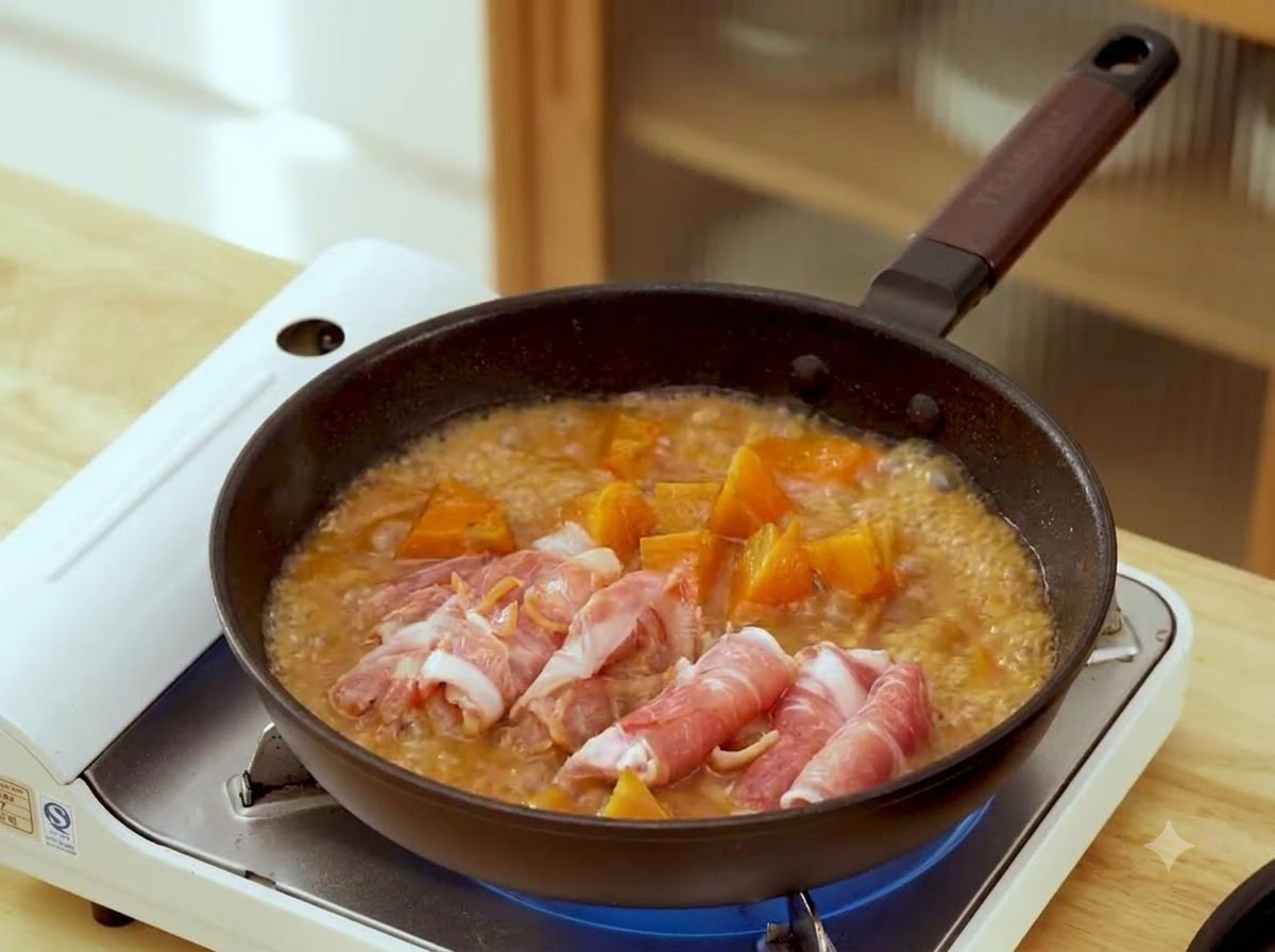 Finas rodajas de ternera cruda enrolladas dispuestas dentro de una sartén con caldo de miso marrón burbujeante y trozos de zanahoria.
