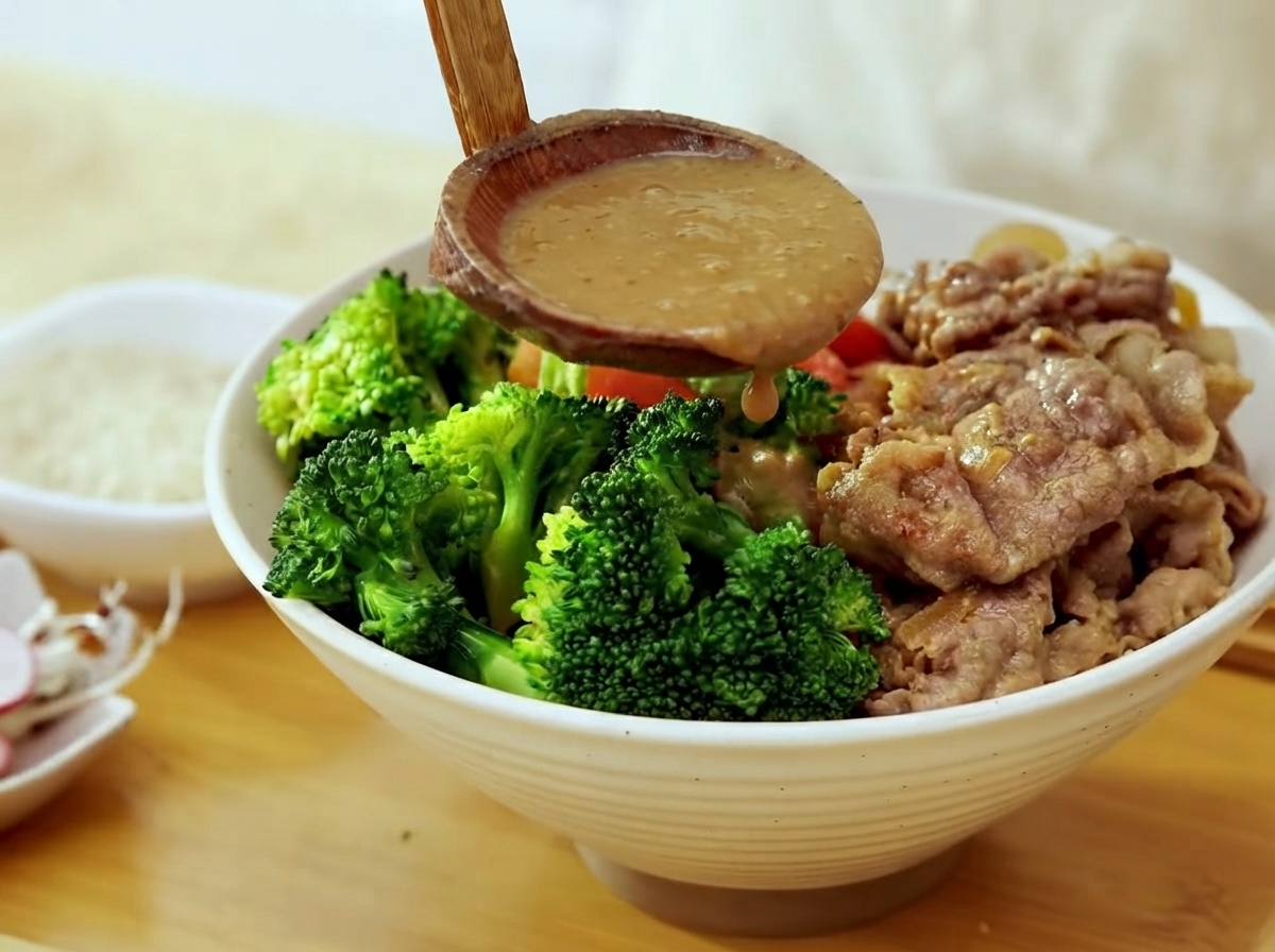 Un cucharón de madera vertiendo salsa de miso marrón espesa sobre un bol lleno de brócoli, ternera cocinada y arroz al vapor.