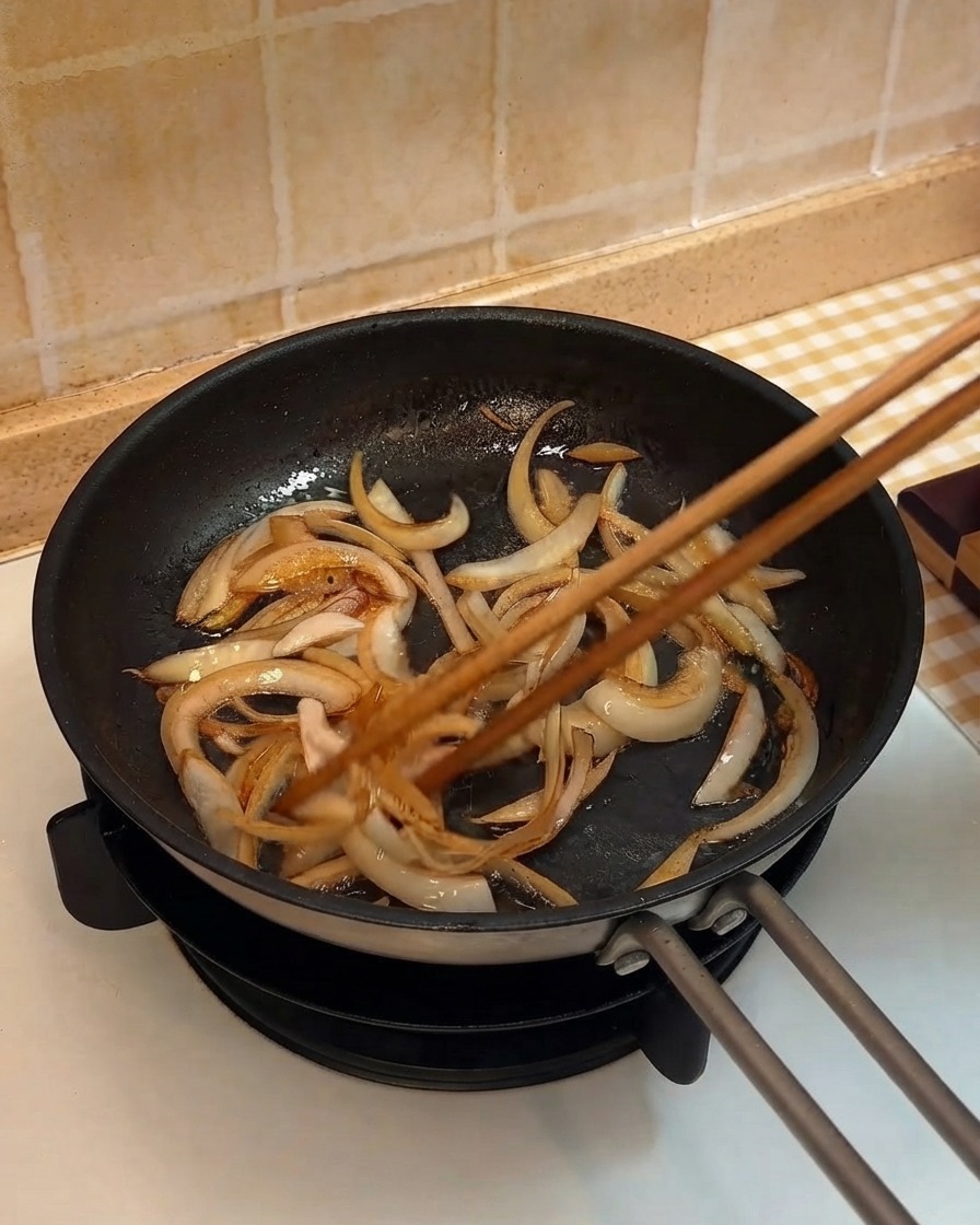 Tiras de cebolla siendo salteadas en una sartén usando palillos de madera.