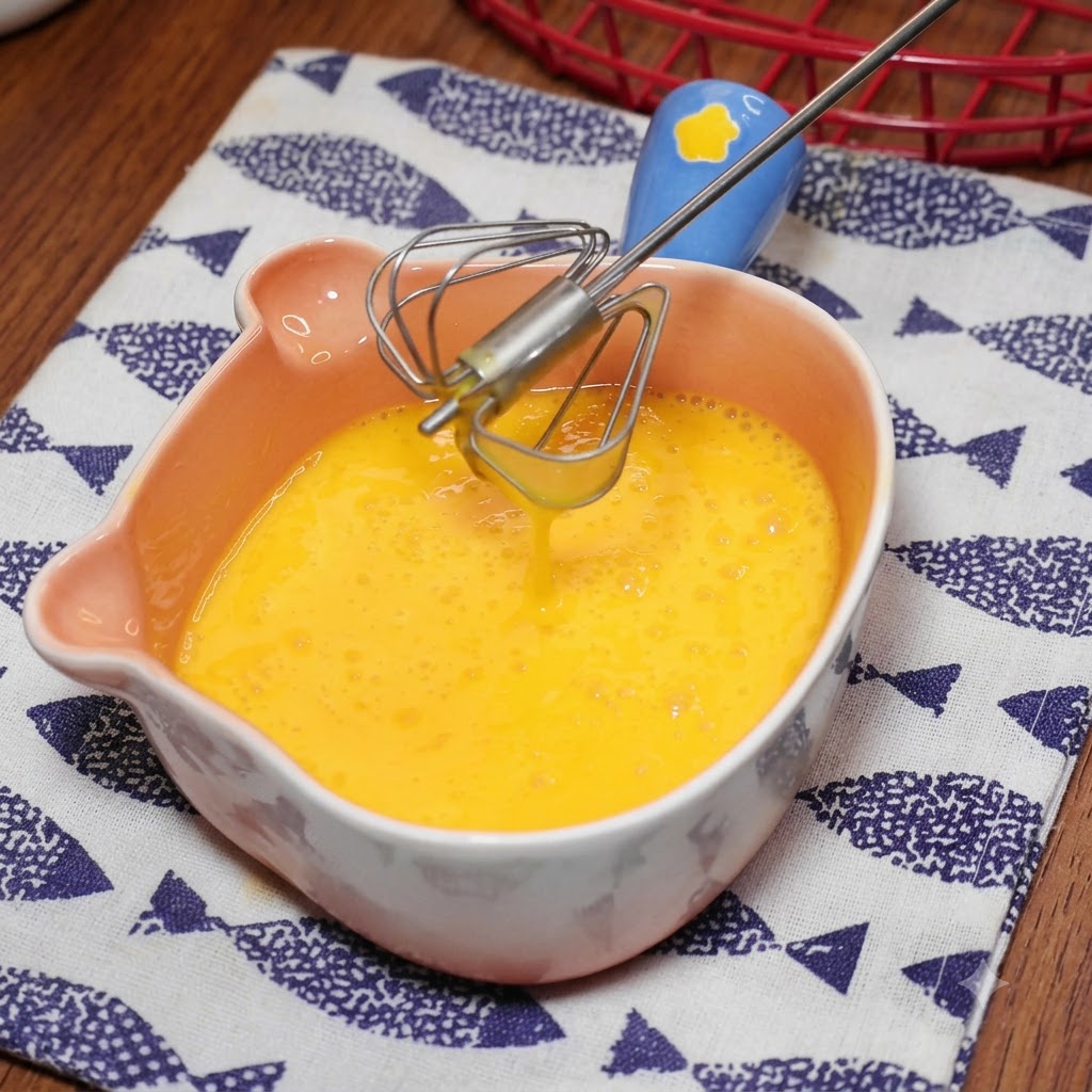 Un pequeño batidor de mango azul mezclando huevos amarillos dentro de un cuenco rosa con forma de oso.