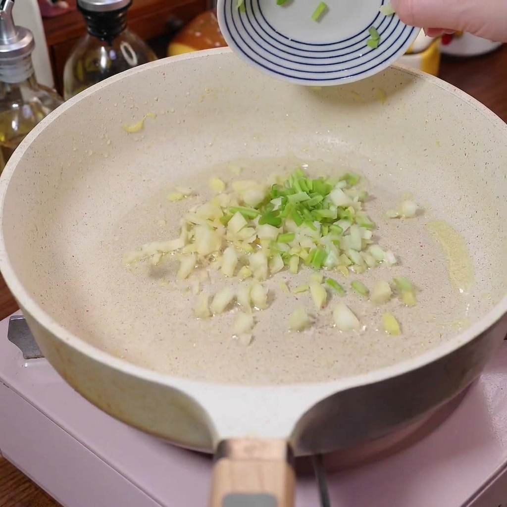 Cebollino picado, ajo y jengibre chisporroteando en una sartén blanca.