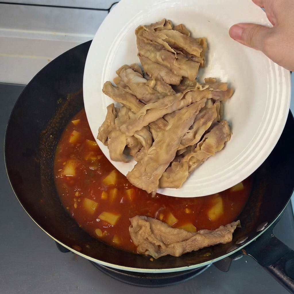 Rollos de carne preblanqueados deslizándose desde un plato blanco hacia el caldo rojo y sabroso en el wok.