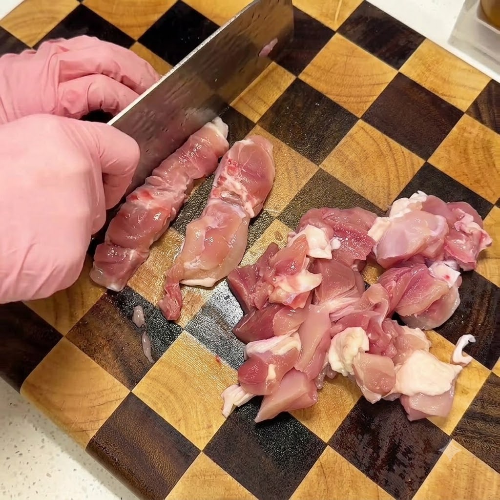 Manos con guantes rosados usando una cuchilla para cortar muslos de pollo crudos en una tabla de cortar de madera a cuadros.