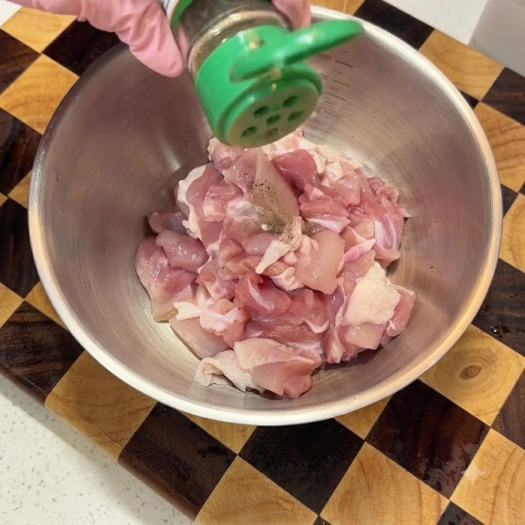 Espolvoreando pimienta negra en polvo desde un frasco verde sobre pollo crudo cortado en cubitos en un bol de metal.